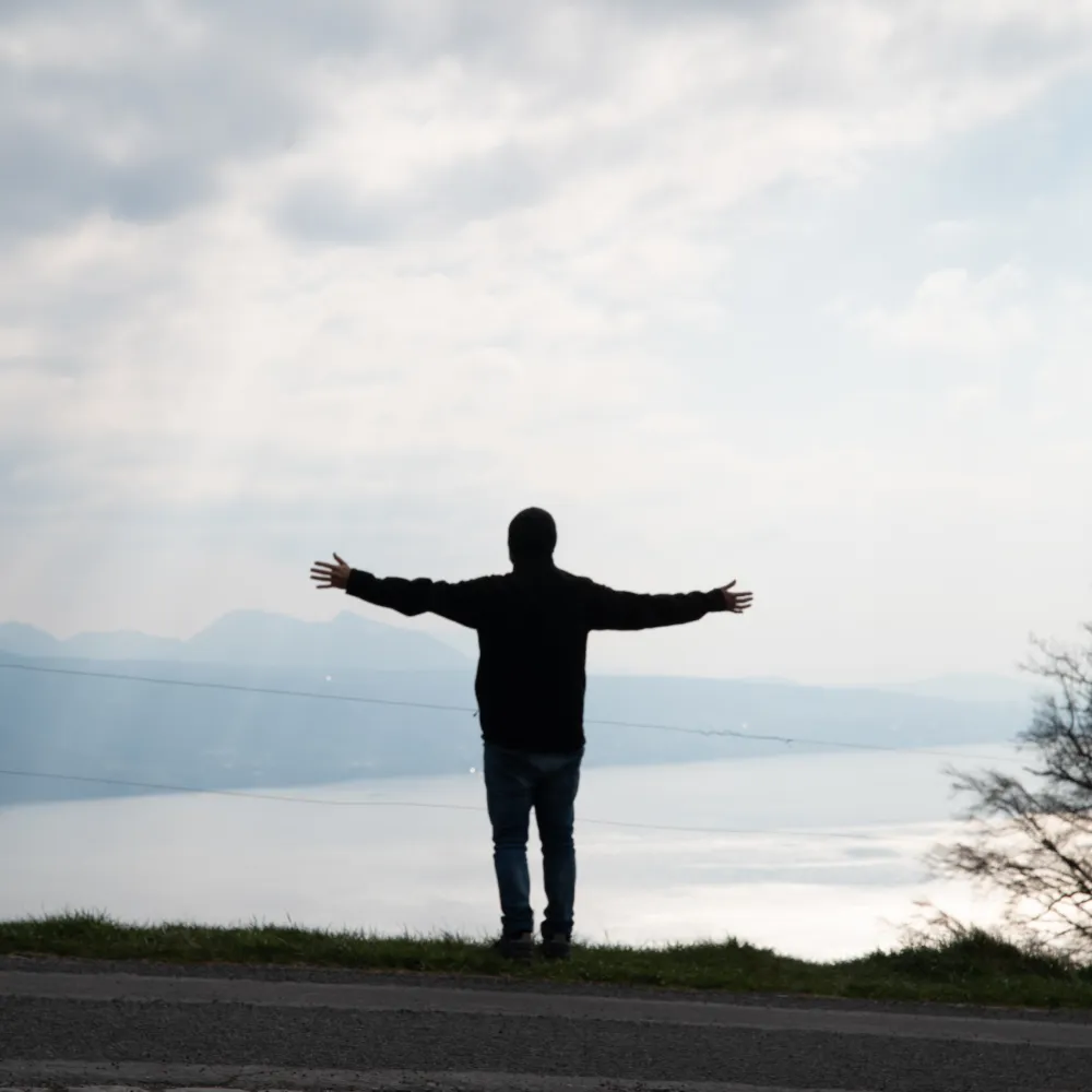 A man standing with his arms spread out overlooking a view of a mountain and a lake.