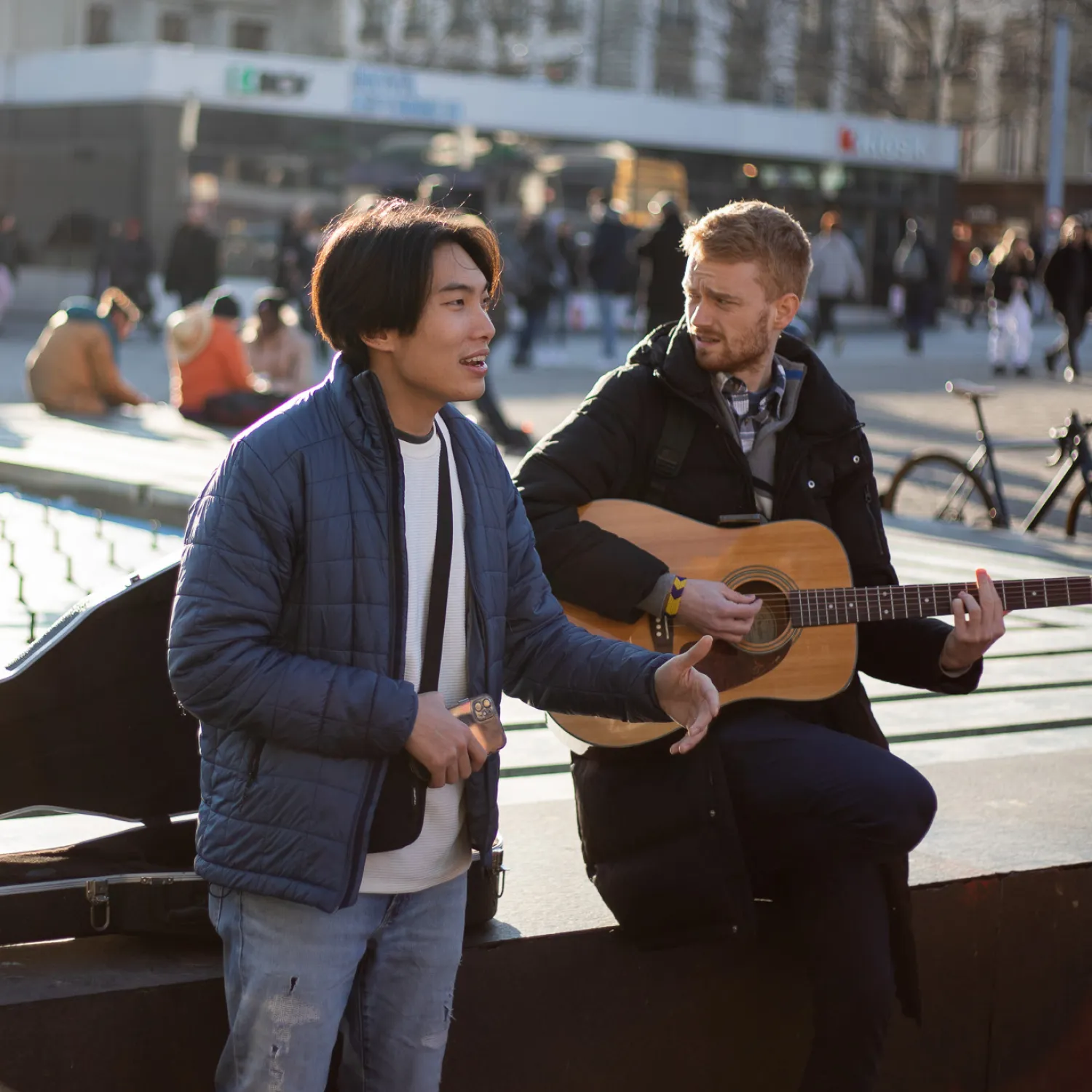 A man playing the guitar next to a man speaking with his arms out.