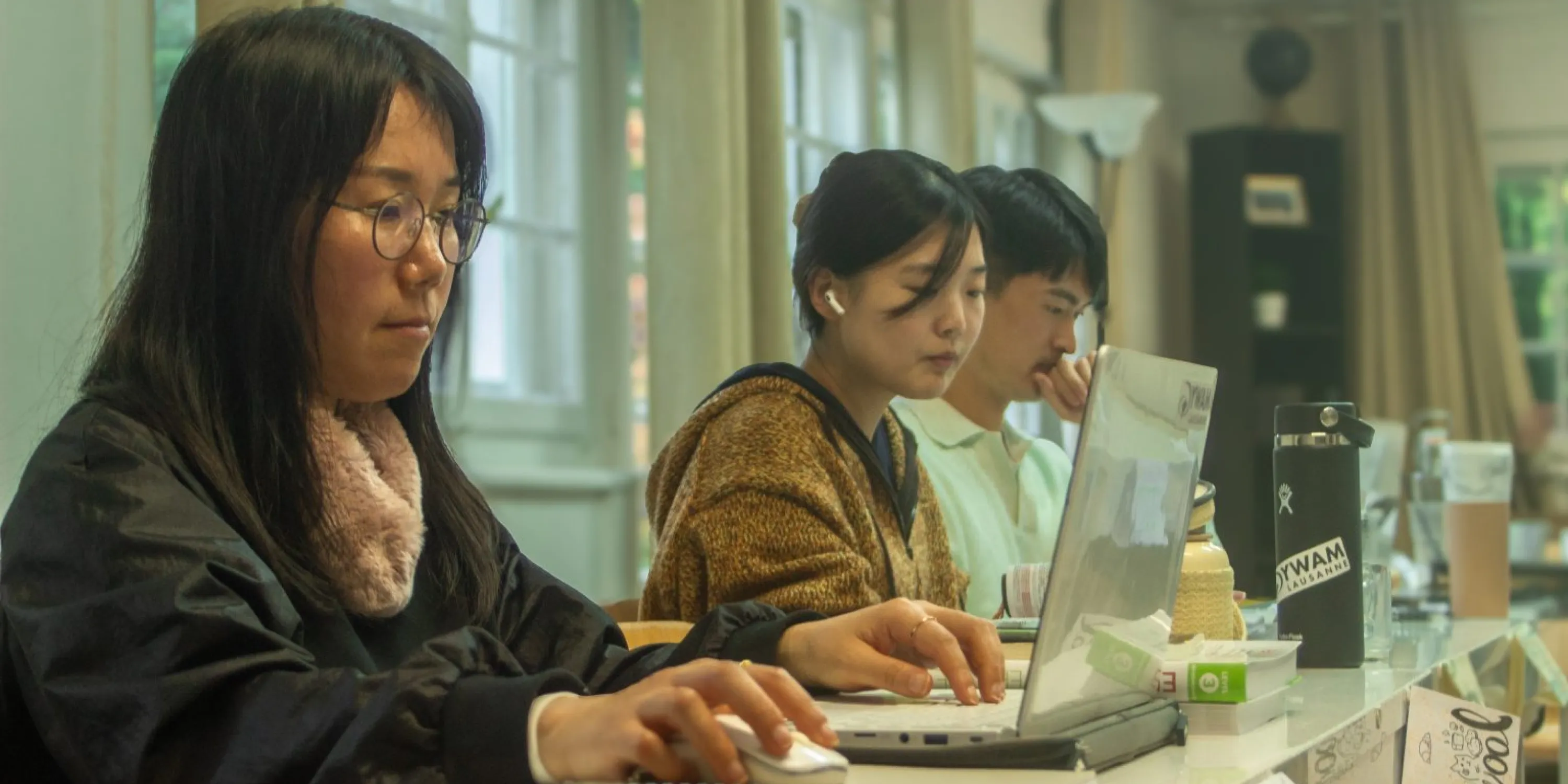 Three students seated at a desk working at their laptops in a classroom.